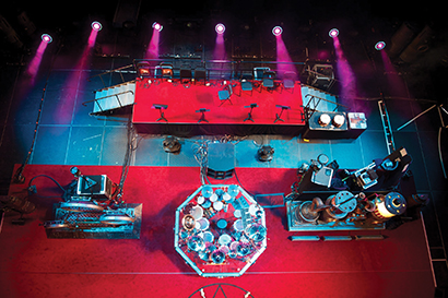 Fig 1: This fly gallery view looking downward onto the Rush stage for the Clockwork Angels tour shows the extremely close proximity of the string section (red platform at top of photo) to the drums, drum fills and instrument amplifiers. The yellow-colored DI boxes are visible along the top edge of the riser.