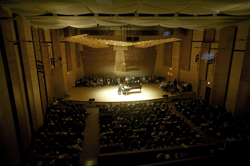 View of the Harris Hall during a solo piano performance. Suspended above the stage is the “mothership” — a large reflective panel that can also house audio and networking electronics, as well as a small central L/R cluster used for pops concerts and other events.