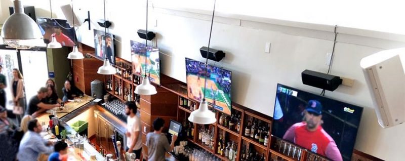 The club is divided into 5 main sections: The Bar, with 20 stools, 6 screens and 5 separate audio zones; two 10-seat Booths, each with their own dedicated screen, club-level sound zones and under-seat subwoofers; a second-level Sky Box with 6 screens and 2 sound zones; a Rivalry Room featuring two dueling pull-out wood bleachers, each with seating for 30, and back to back projector screens in the center of the room that drop down between the bleachers for each team’s fans to watch from the other side; and a 150-person private event space with 12 screens and a 160” projector.